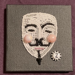 May include: A beaded mask design, featuring a white face with black details and pink cheeks, is mounted on a gray textured square. A small, round, white and black button with the letter "Z" is next to the mask. The mask is a symbol of anonymity.