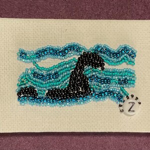 May include: A rectangular piece of off-white canvas featuring a beaded design of a black sea creature swimming through wavy lines of light blue, turquoise, and dark blue seed beads. A single white circular bead with the letter Z is attached.