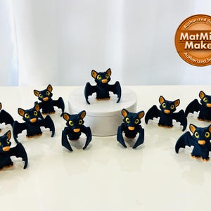 May include: Ten black bat figurines with orange eyes and wings spread out. The bats are arranged in a circle on a white surface. The bats are about 2 inches tall.