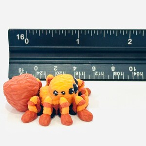 May include: A handmade orange and yellow spider figurine with black accents, next to a ruler. The spider has eight legs and a round body. The ruler shows measurements in centimetres.