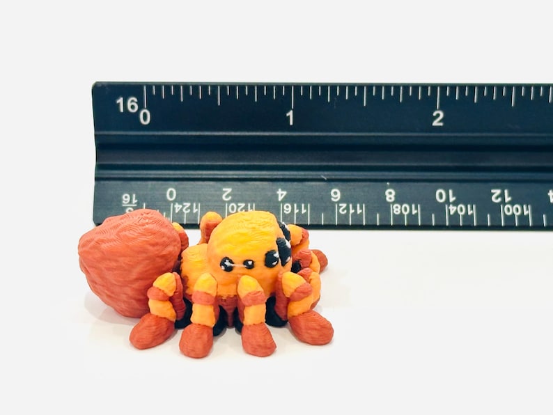 May include: A handmade orange and yellow spider figurine with black accents, next to a ruler. The spider has eight legs and a round body. The ruler shows measurements in centimetres.