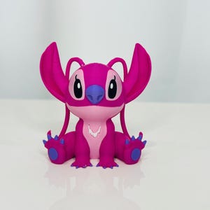 May include: A pink and purple plush toy of a cartoon character with large ears and a wide smile. The character is sitting with its legs crossed and its arms resting on its lap.