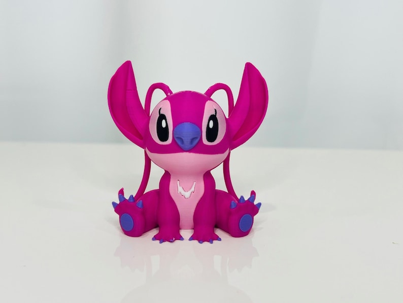 May include: A pink and purple plush toy of a cartoon character with large ears and a wide smile. The character is sitting with its legs crossed and its arms resting on its lap.