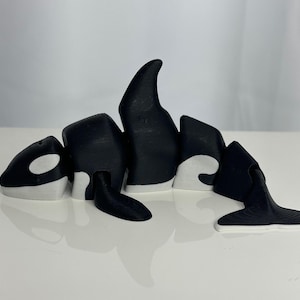 May include: A black and white 3D printed orca figurine. The orca is posed swimming with its dorsal fin raised. The orca has a white patch around its eye and a white underside.