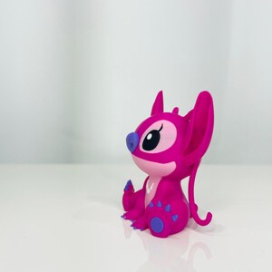 May include: A pink Stitch figurine with purple accents. The figurine is sitting on a white surface.