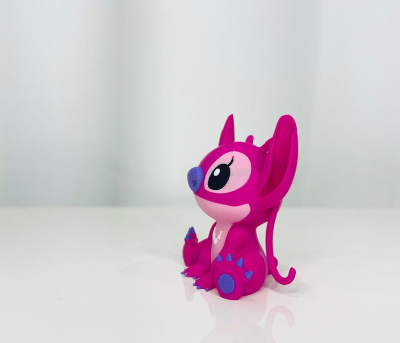 May include: A pink Stitch figurine with purple accents. The figurine is sitting on a white surface.