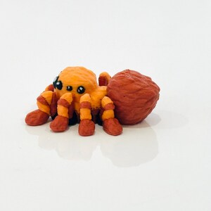 May include: A handcrafted orange and red spider ornament with black eyes. The spider has a textured body and legs with alternating orange and red stripes. The ornament is set against a white background.