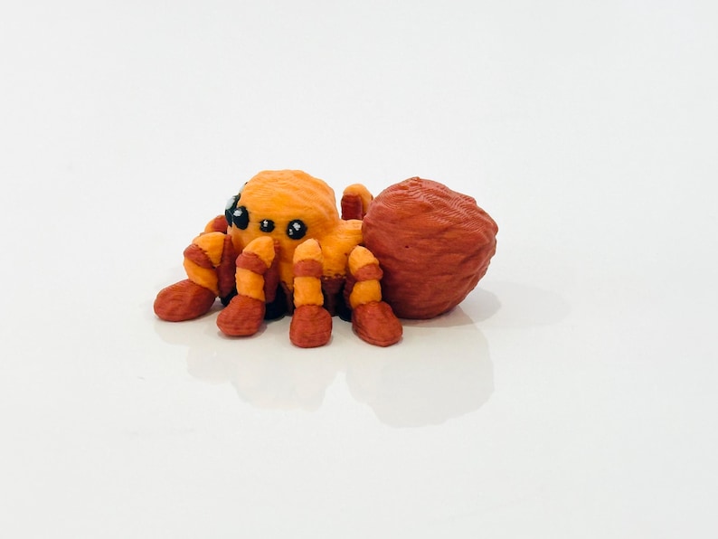 May include: A handcrafted orange and red spider ornament with black eyes. The spider has a textured body and legs with alternating orange and red stripes. The ornament is set against a white background.