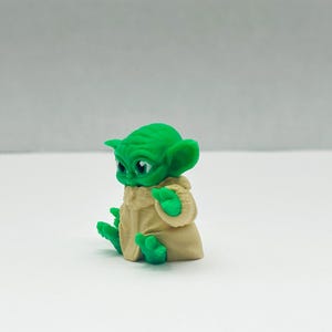 Puede incluir: Peque&ntilde;a figura verde de un personaje con orejas grandes y una t&uacute;nica beige. La figura est&aacute; sentada con las manos levantadas y los ojos mirando hacia delante. La figura est&aacute; sobre una superficie blanca.