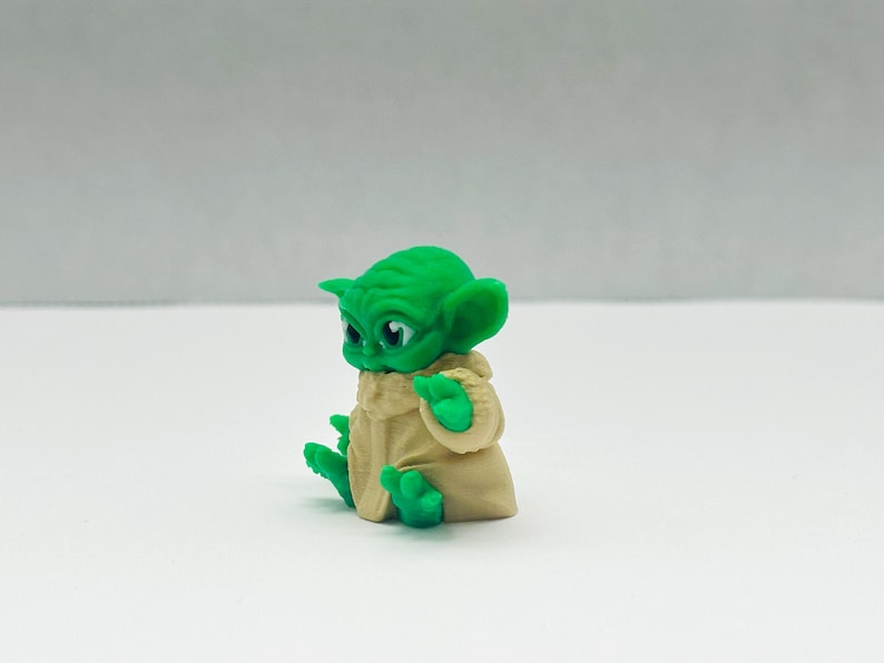 Puede incluir: Peque&ntilde;a figura verde de un personaje con orejas grandes y una t&uacute;nica beige. La figura est&aacute; sentada con las manos levantadas y los ojos mirando hacia delante. La figura est&aacute; sobre una superficie blanca.