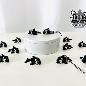 May include: A collection of black and white orca-shaped charms, some with attached cords, are scattered on a white surface. Several charms are arranged around a white, cylindrical display stand. The image includes a logo that reads "3DGOB AUTHORIZED SELLER."