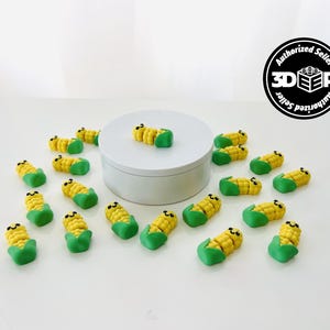 May include: A collection of miniature corn-shaped figurines. Each figurine features yellow corn kernels, green husks, and small black eyes, arranged on a white surface. A white circular platform is in the center.