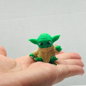 Puede incluir: Peque&ntilde;a figura verde de un personaje con orejas y ojos grandes, vestido con una t&uacute;nica beige. La figura descansa en la palma de una mano. El personaje es de la franquicia Star Wars.