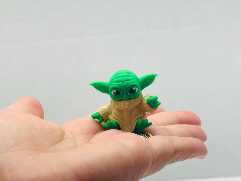 Puede incluir: Peque&ntilde;a figura verde de un personaje con orejas y ojos grandes, vestido con una t&uacute;nica beige. La figura descansa en la palma de una mano. El personaje es de la franquicia Star Wars.