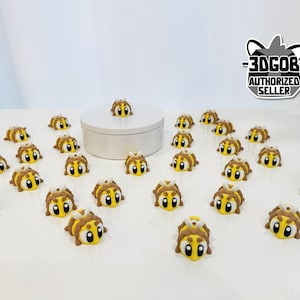 May include: A collection of small, yellow and brown bee-shaped figurines with large, cartoonish eyes. The figurines are scattered on a white surface, with one placed on a small, round, white platform. The image includes a logo that says "-3DCOB- AUTHORIZED SELLER."