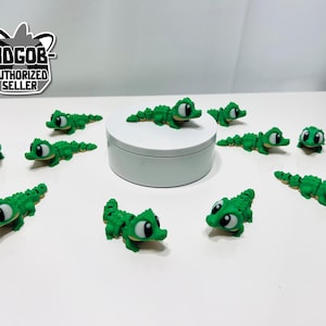 May include: A collection of small, green alligator figurines with large, cartoonish eyes. The alligators are scattered around a white platform. The image includes a logo that says "3DGOB AUTHORIZED SELLER".
