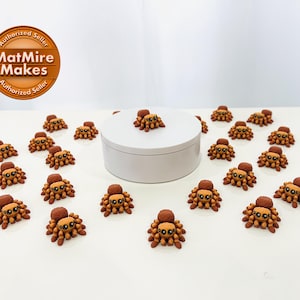 May include: A collection of miniature spider figurines, each with a brown body, lighter brown abdomen, and a dark brown head. The spiders are arranged around a white, circular platform. The image includes a logo.