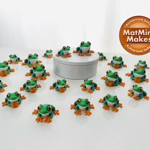 May include: A collection of small, green and blue frog figurines with orange feet and a white belly. The frogs are arranged on a white surface, some on a small white platform. The image includes a circular logo that says "MatMire Makes".