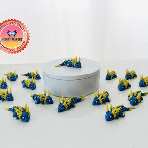 May include: A collection of blue and yellow dragon figurines. The dragons are arranged on a white surface, some around a white cylinder. The figurines have yellow spikes and accents. The image includes a "Mighty Makers" authorized seller sticker.