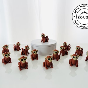 May include: A collection of small, brown monkey figurines with tan faces and large, round eyes. One monkey sits atop a white pedestal, while the others are arranged on a white surface. The image includes a circular "AUTHORIZED SELLER" logo.