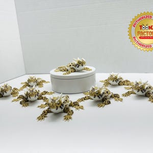 May include: A collection of eight small, white and brown, horned lizard figurines. The figurines are arranged in a circle around a white ceramic box. The box has a lid and is decorated with a gold sticker that says "ToonZ Factory".