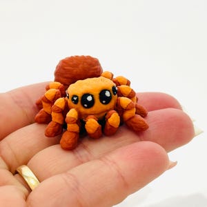 May include: A handmade, orange and brown spider figurine with large, expressive eyes. The spider has a rounded body and segmented legs, and is likely made of clay or a similar material. The figurine is held in a hand.