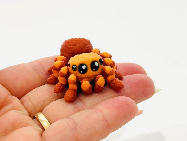May include: A handmade, orange and brown spider figurine with large, expressive eyes. The spider has a rounded body and segmented legs, and is likely made of clay or a similar material. The figurine is held in a hand.