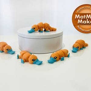 May include: Five orange and blue platypus figurines. The platypus are posed in a variety of positions. The figurines are on a white surface. The image also includes a round, white container and a brown circle with the text "MatMire Makes Authorized Seller".