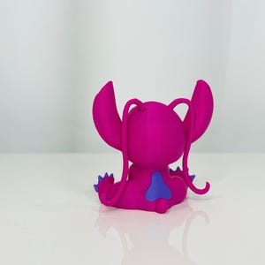 May include: A pink and purple Stitch figurine with a blue heart on its back. The figurine is sitting on a white surface.
