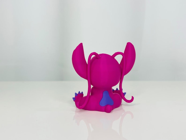 May include: A pink and purple Stitch figurine with a blue heart on its back. The figurine is sitting on a white surface.