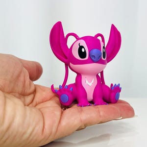 May include: A pink and purple Stitch figurine with large ears and a white necklace. The figurine is sitting on a hand.