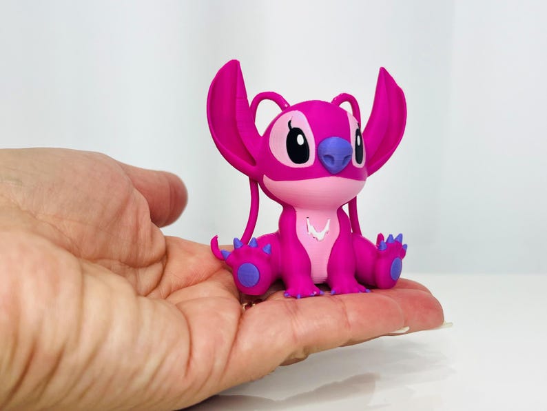May include: A pink and purple Stitch figurine with large ears and a white necklace. The figurine is sitting on a hand.