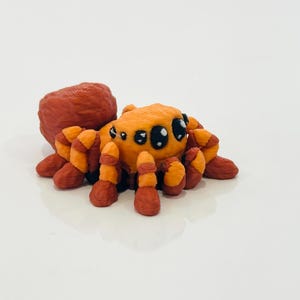 May include: A handmade orange and red spider figurine with large black eyes. The spider has segmented legs and a rounded body, and is likely made of clay or a similar material. The figurine is set against a white background.