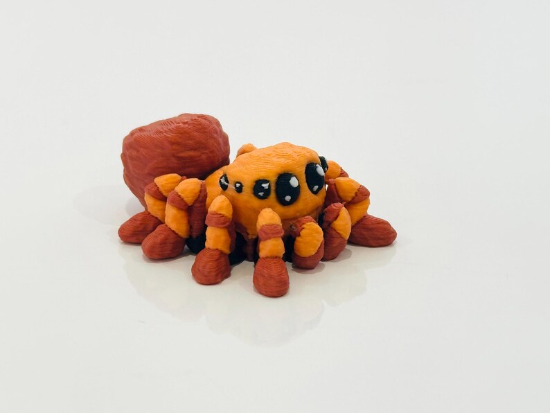 May include: A handmade orange and red spider figurine with large black eyes. The spider has segmented legs and a rounded body, and is likely made of clay or a similar material. The figurine is set against a white background.
