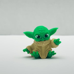 Puede incluir: Una figura verde de un personaje con grandes orejas y ojos, vistiendo una t&uacute;nica beige. La figura est&aacute; sentada con las manos extendidas. El personaje es de una popular franquicia de ciencia ficci&oacute;n.