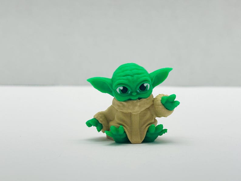 Puede incluir: Una figura verde de un personaje con grandes orejas y ojos, vistiendo una t&uacute;nica beige. La figura est&aacute; sentada con las manos extendidas. El personaje es de una popular franquicia de ciencia ficci&oacute;n.