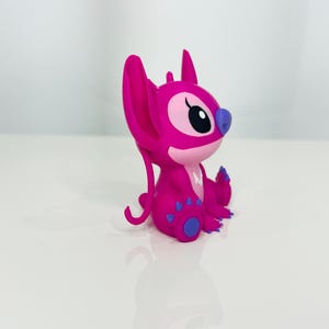 May include: A pink Stitch figurine with purple accents. The figurine is sitting with its legs crossed and its arms folded.
