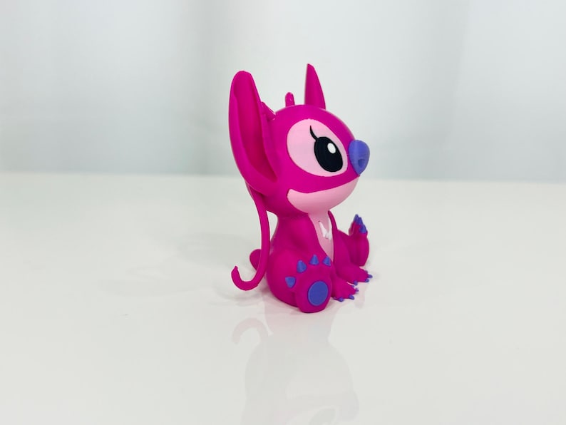 May include: A pink Stitch figurine with purple accents. The figurine is sitting with its legs crossed and its arms folded.