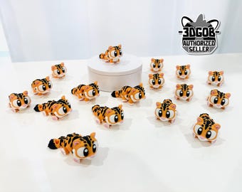 May include: A collection of orange and black tiger figurines with large eyes and white paws. The figurines are arranged on a white surface, some standing and some lying down. A white cylinder is in the background.