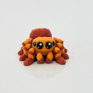 May include: A small, orange and brown spider figurine with large, cartoonish eyes. The spider has a rounded body and eight legs, all in shades of orange and brown. The figurine is set against a plain white background.