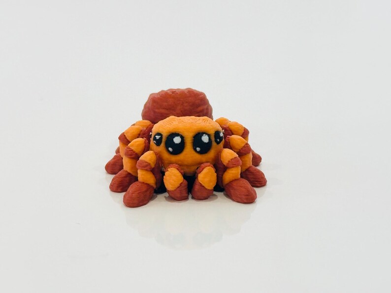 May include: A small, orange and brown spider figurine with large, cartoonish eyes. The spider has a rounded body and eight legs, all in shades of orange and brown. The figurine is set against a plain white background.