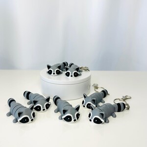 May include: Six gray raccoon keychain charms with black eyes and a white face. The charms are on a white surface with a white round container in the background.