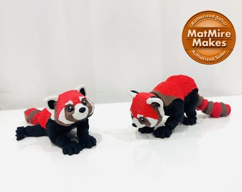Puede incluir: Dos figuritas de panda rojo, una tumbada y la otra de pie, ambas con marcas negras y blancas. Las figuritas están hechas de un material suave y de felpa.