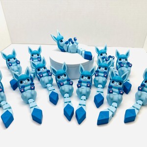 May include: A collection of light blue and dark blue articulated figurines, resembling a cartoon creature with large ears and a segmented tail. One figurine rests on a white platform, while others are arranged around it. The figurines have dark blue accents and paw-like hands.
