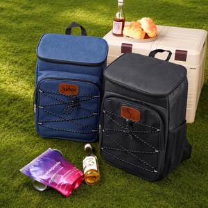 May include: Two rectangular insulated backpacks, one blue and one black, each featuring a front pocket with a brown leather-like patch, a black bungee cord lacing system, and a top handle. The blue patch displays the name Aiden, while the black patch shows the letters ST.