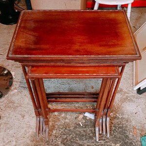 Puede incluir: Un juego de tres mesas nido de madera con un acabado en tono marrón rojizo intenso. Cada mesa rectangular presenta una superficie lisa con un patrón de borde incrustado decorativo y patas rectas y delgadas unidas por soportes horizontales.