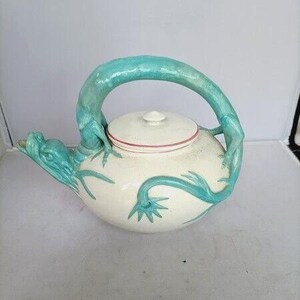 May include: A cream-colored ceramic teapot featuring a sculpted teal dragon that forms the handle and spout. The dragon's body wraps around the side of the pot, and the lid is accented with a thin red line around the rim, creating a distinct contrast.