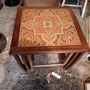 May include: A square wooden nesting table featuring a tabletop inlaid with four ceramic tiles. The tiles display a symmetrical, intricate brown and cream geometric pattern. The table has a dark wood frame with curved legs and a horizontal support bar.