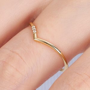 May include: A thin gold V-shaped ring featuring a polished band and three small, clear, round-cut stones set at the center point of the chevron design. The ring has a smooth, metallic finish and a delicate, minimalist structure that follows a V-shape.