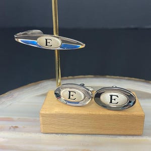 Puede incluir: Un conjunto de accesorios de metal plateado que incluye un pasador de corbata y dos gemelos ovalados. Cada pieza presenta una incrustación central de plata cepillada con una letra E mayúscula negra grabada, rodeada por un borde plateado pulido.